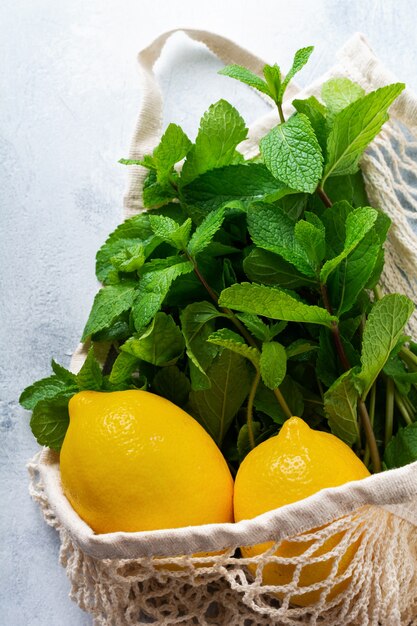 Hojas de menta, limón fresco y miel acostada sosteniendo una bolsa de algodón de compras de malla sobre hormigón gris viejo.