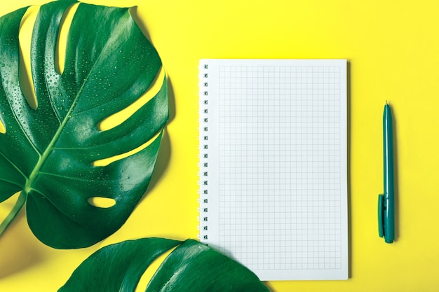 Hojas de Monstera, un cuaderno a cuadros y un bolígrafo sobre un moderno fondo amarillo. Copia espacio