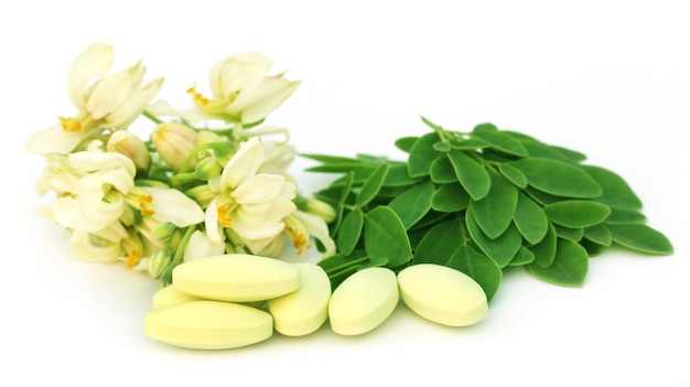 Hojas de moringa y flores con pastillas sobre fondo blanco.