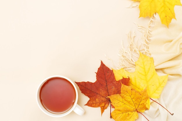 Hojas de otoño con una taza de té o café junto a una bufanda sobre un fondo beige con espacio para copiar