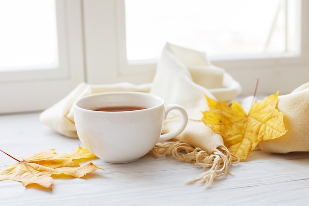 Hojas de otoño con una taza de té o café junto a una bufanda sobre un fondo de madera con espacio para copiar