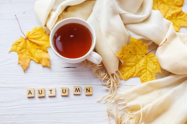 Hojas de otoño con una taza de té o café junto a una bufanda sobre un fondo de madera con espacio para copiar