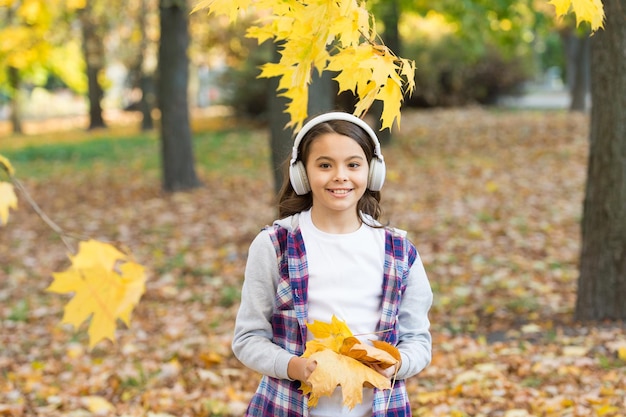 Las hojas que caen una niña feliz usa auriculares en el paisaje de otoño una sonrisa de niño linda con auriculares estéreo un niño pequeño escuchando auriculares modernos tecnología de auriculares melodía del otoño