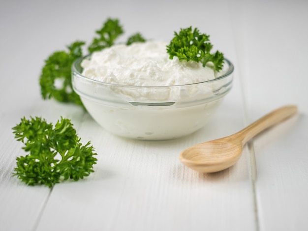 Hojas rizadas de perejil y un tazón de crema cuajada y cuchara sobre una mesa blanca.