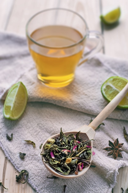 Hojas de té verde en una cuchara de madera sobre textiles de lino con rodajas de limón y taza de té, cerrar