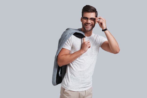 Foto ¡hola! apuesto joven ajustando sus gafas y sonriendo mientras está de pie contra el fondo gris