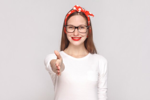 Hola, encantado de conocerte. apretón de manos retrato de hermosa mujer joven emocional en camiseta blanca con pecas, gafas negras, labios rojos y banda para la cabeza. Foto de estudio en interiores, aislado sobre fondo gris claro