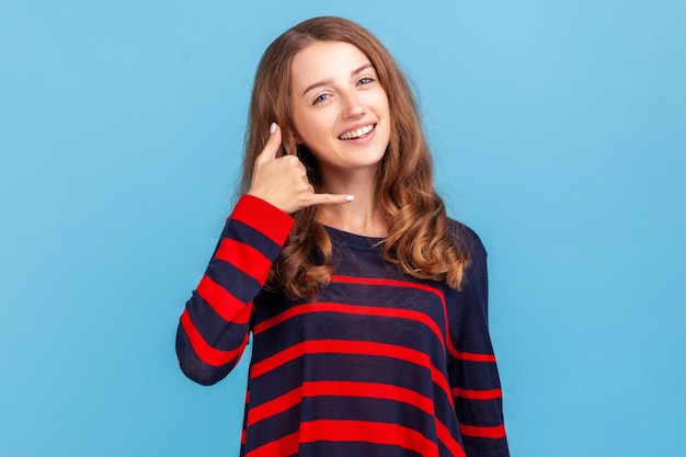 Hola, guapo, llámame Mujer sonriente con un suéter de estilo informal a rayas coqueteando sosteniendo los dedos cerca de la oreja mostrando un gesto de llamada respondiendo una llamada Estudio interior aislado en fondo azul