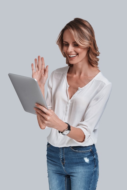 ¡Hola! Hermosa mujer joven mirando la mesa digital y saludando con una sonrisa en su rostro mientras está de pie contra el fondo gris