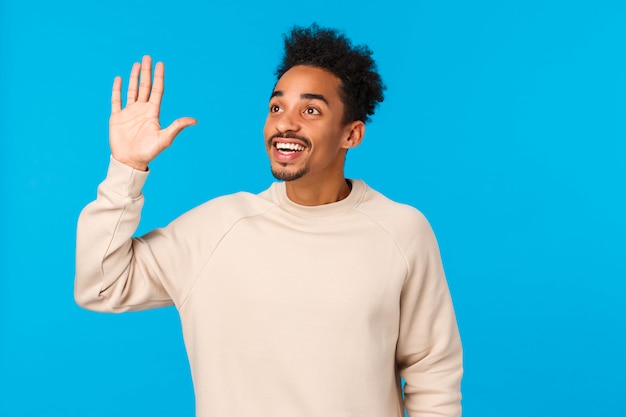 Hola hola, por aquí. Atractivo amistoso hipster afroamericano chico corte de pelo afro, bigote, ver a un amigo en la multitud, mirando la esquina superior izquierda, saludando con la mano en gesto, sonriendo, fondo azul