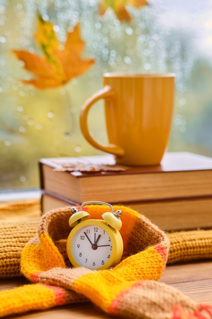 Hola otoño. Despertador amarillo envuelto en una bufanda, taza de libros cerca de la ventana, está lloviendo