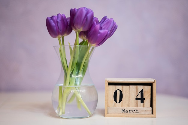 Hola primavera Calendario cúbico de madera con fecha del 4 de marzo rodeado de ramo de tulipanes morados con hojas verdes en jarrón de vidrio