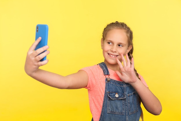 Hola. Retrato de una adorable niñita con overoles de mezclilla saludando mientras se comunica con sus padres en una videollamada, tomándose selfie en el teléfono móvil. tiro de estudio interior aislado sobre fondo amarillo