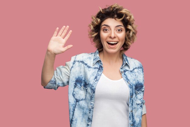 Hola. Retrato de mujer joven feliz sorprendida con peinado rizado en camisa azul casual de pie, saludando, agitando la mano y mirando con una gran sonrisa. Foto de estudio de interior, aislado sobre fondo rosa