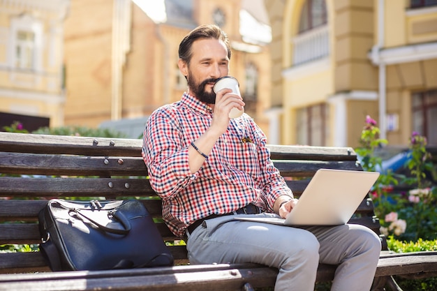 Holen Sie sich etwas Energie. Netter bärtiger Mann, der Kaffee trinkt, während Laptop auf der Bank benutzt