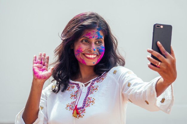 Holi Festival Of Colors Retrato de niña india feliz en color holi ella tomando una foto selfie en la cámara del teléfono inteligente en el fondo del estudio de pared blanca