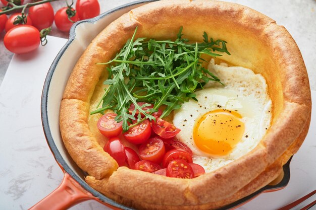 Holländischer Baby-Pfannkuchen Frischer hausgemachter holländischer Baby-Pfannkuchen mit Spiegeleier-Tomaten und grünem Rucola in Reed-Gusseisenpfanne auf hellgrauem Betontisch im Landhausstil Flach lag in der Nähe