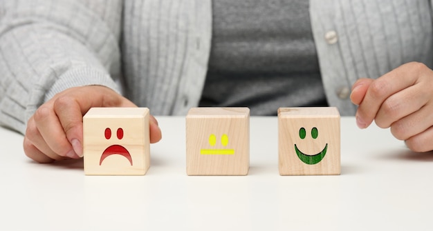 Holzblöcke mit unterschiedlichen Emotionen von Lächeln bis Traurigkeit und die Hand einer Frau. Konzept zur Beurteilung der Qualität eines Produkts oder einer Dienstleistung, emotionaler Zustand, Nutzerbewertungen