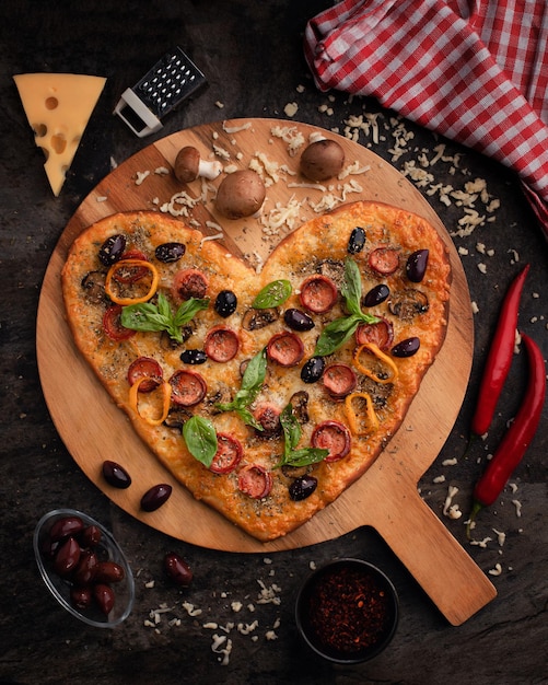 Holzbrett mit leckerer herzförmiger Pizza mit Zutaten auf schwarzem Hintergrund Valentinstagsfeier