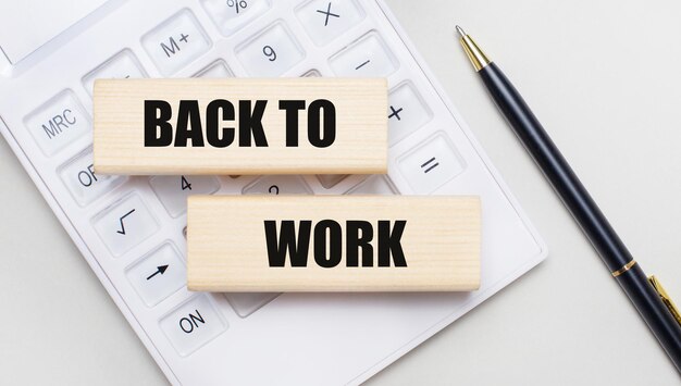 Holzklötze mit dem BACK TO WORK Iie auf hellem Hintergrund auf einem weißen Taschenrechner In der Nähe befindet sich ein schwarzer Griff Geschäftskonzept