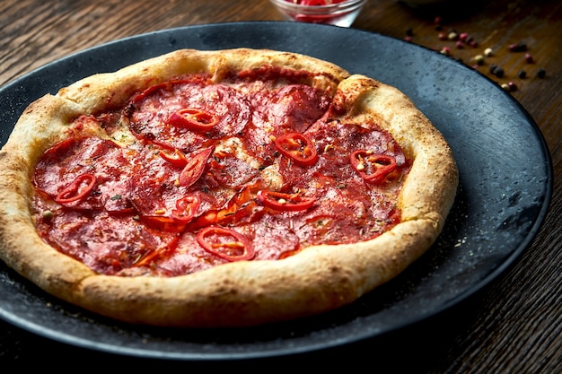 Holzofenpizza mit würziger Chorizo-Salami und Chili mit knusprigen Beilagen, serviert auf einem schwarzen Teller auf einem Holztisch. Pizzette eine Art italienische Pizza