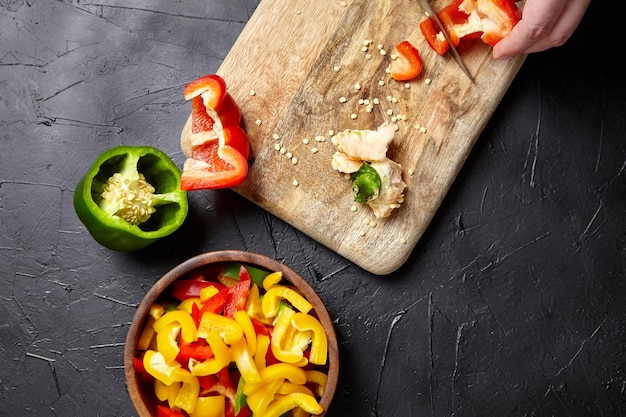 Holzschneidebrett und Schüssel mit Stücken verschiedener Paprika auf schwarzem Hintergrund, Draufsicht