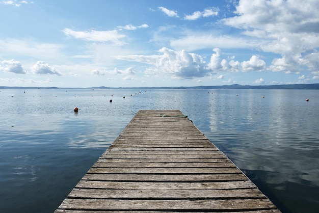 Holzsteg für Boote vor blauem Himmel mit einigen Wolken Schwimmende Bojen Hügel im Hintergrund Bolsenasee Italien