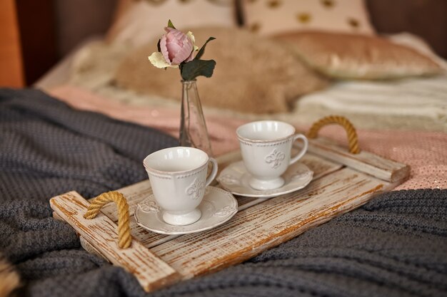 Holztablett des Kaffees mit Blume auf Bett. Romantisches Bett mit verschiedenen warmen Decken und Kissen. Bleib zu Hause, Quarantäne.