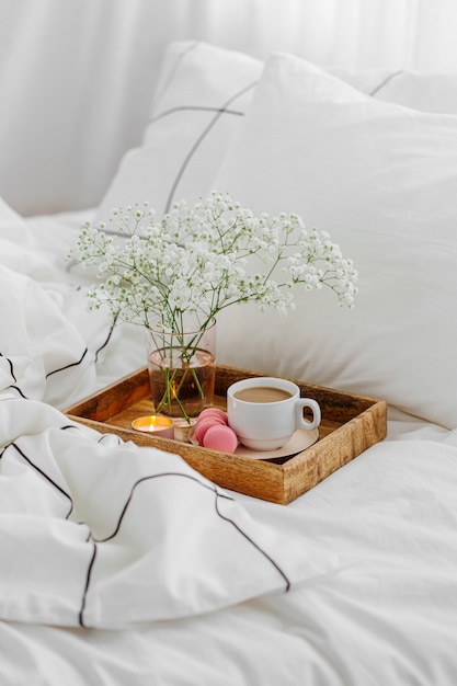 Holztablett Kaffee und Kerzen mit Blumen auf dem Bett. Weiße Bettwäsche mit gestreifter Decke und Kissen. Frühstück im Bett. Hygge-Konzept.