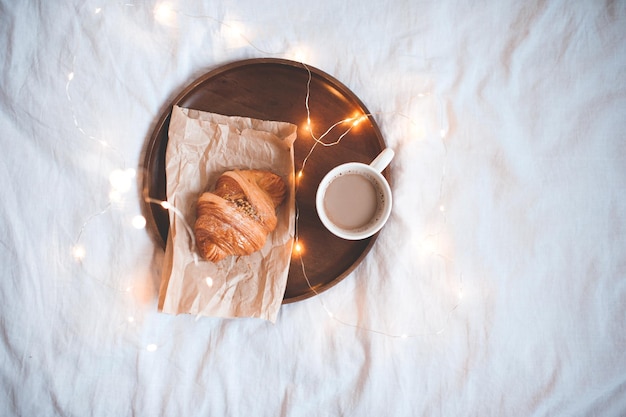 Holztablett mit leckerem Frühstück: Freah Croissant und Tasse Kaffee im Bett auf weißer Bettwäsche.