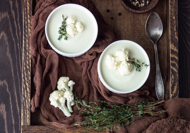 Holztablett mit zwei Keramikschalen mit Blumenkohlcremesuppe garnieren mit frischem Blumenkohl, Thymian und Brot. Herbst- oder Winterkost.