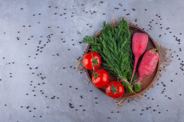 Holzteller mit rotem Rettich, Tomaten und Dill auf Steinhintergrund.