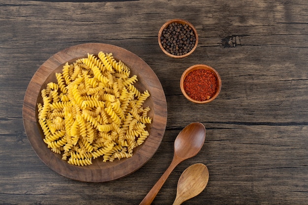 Holzteller mit ungekochten Fusilli-Nudeln und Gewürzen auf Holzoberfläche.