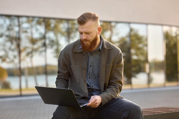 Un hombre de 35 años con una barba roja se sienta en un banco con una computadora portátil durante la pausa para el almuerzo