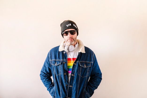 Hombre de 45 años vestido con camiseta blanca con la bandera del orgullo, chaqueta vaquera y sombrero.