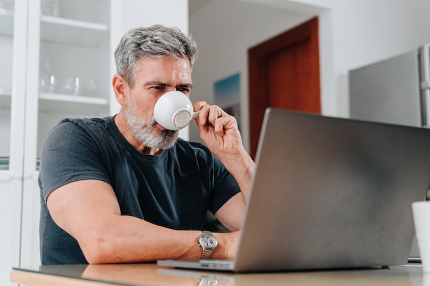 Hombre de unos 50 años que trabaja desde casa mientras toma un café y está en una videollamada en la computadora