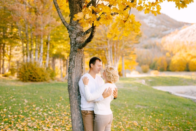 El hombre abraza a la mujer por los hombros cerca de un árbol en el bosque de otoño