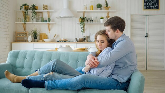 El hombre abraza a la señora sentada en el sofá y besándose en la cocina.