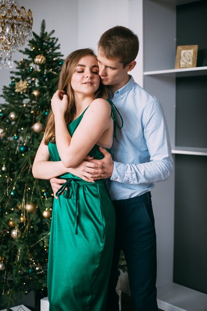 Hombre abrazando a mujer en vestido de noche elegante verde cerca del árbol de Navidad, año nuevo