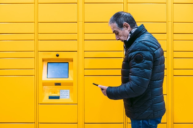 El hombre con abrigo gris mira su celular para recoger un paquete del casillero amarillo. Concepto de mensajería, comparación en línea, comercio electrónico y paquetes.