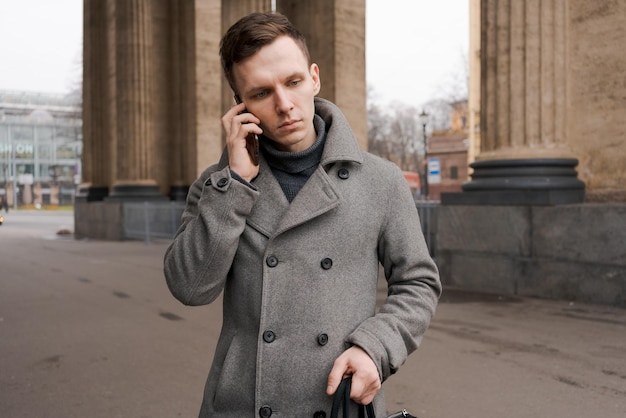 Hombre con abrigo gris y suéter se para en la calle de la ciudad Habla por teléfono Resuelve