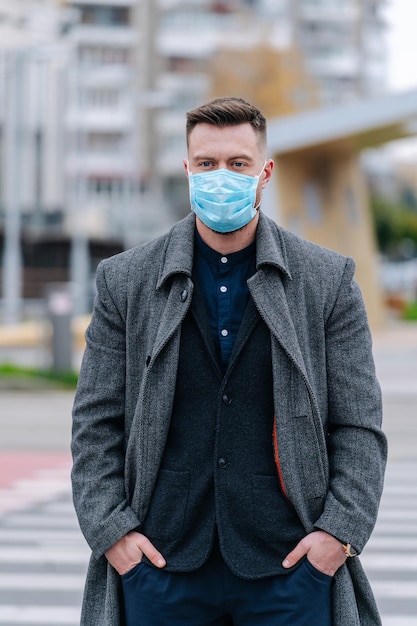 Hombre con abrigo gris y traje oscuro se encuentra en el centro de la ciudad con las manos en los bolsillos. Joven empresario usa máscara facial. De cerca.