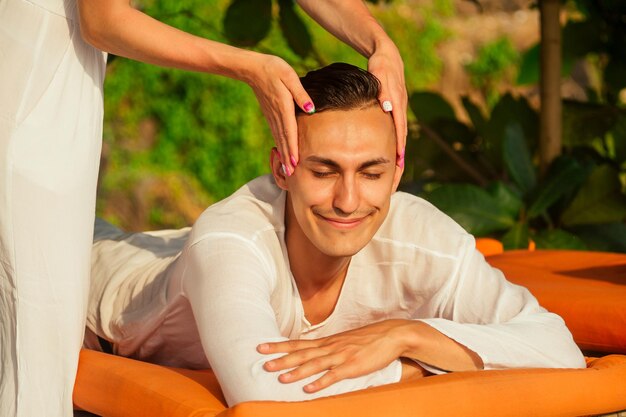 Hombre con aceite de tratamiento de spa Ayurvedahermoso modelo masculino de pelo oscuro barbudo relajándose en la playa y el concepto de relajación plumeria y coco