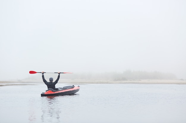 Foto hombre acolchado en acolchado de río, sentado en canoa con remo, disfrutando de los deportes acuáticos y la hermosa naturaleza, vestido con chaqueta negra y gorra gris