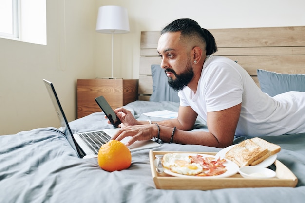 Hombre acostado en la cama, desayunando, trabajando en la computadora portátil y revisando las redes sociales en el teléfono inteligente