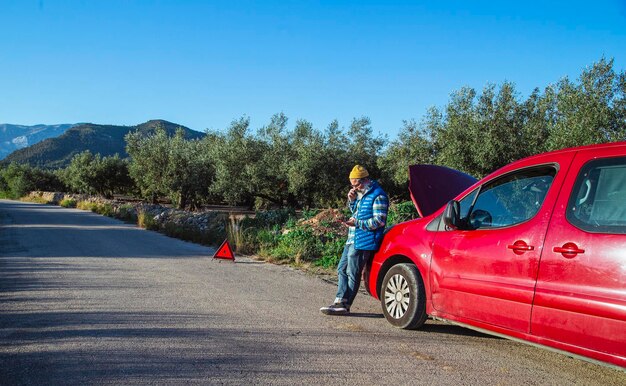 Hombre con actitud relajada varado en la carretera al lado del coche averiado hablando por teléfono persona