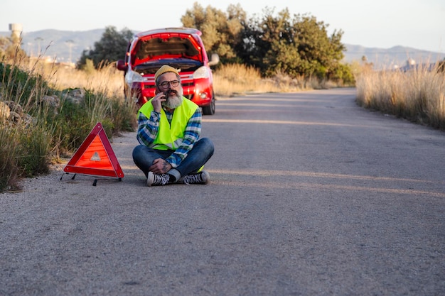 Foto hombre con actitud relajada varado en la carretera junto al coche averiado hablando por teléfono