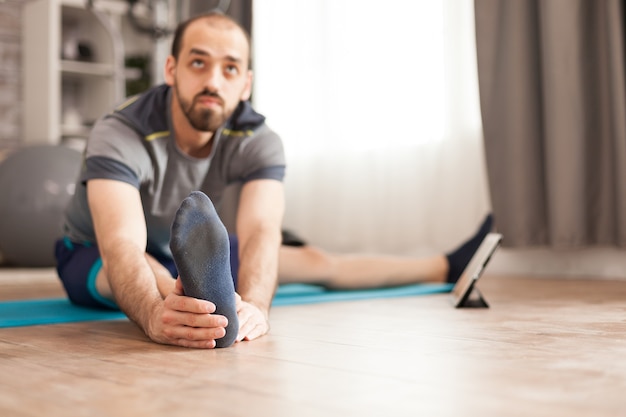 Hombre activo en estera de yoga en casa estirando su cuerpo durante la clase en línea en la computadora de la tableta.