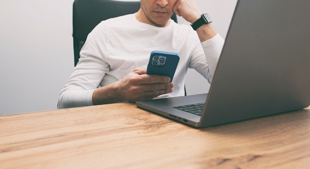 Hombre adicto a su teléfono navegando por las redes sociales en su teléfono mientras debe trabajar con su computadora portátil