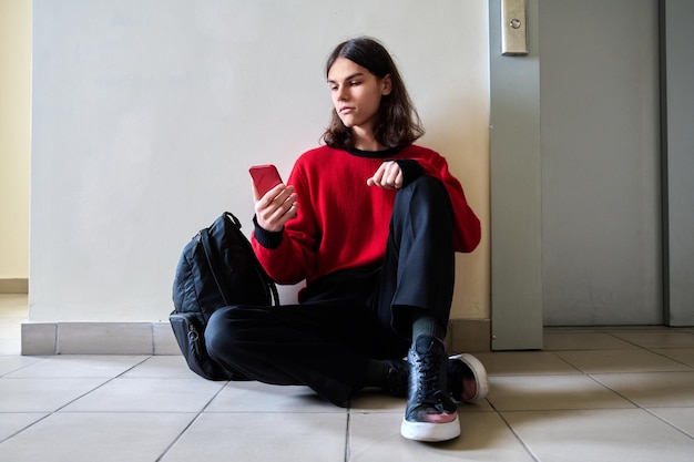 Hombre adolescente de moda con mochila para smartphone sentada en el suelo